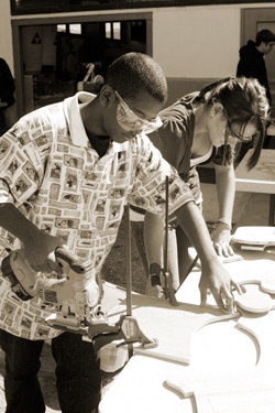 A person wearing safety goggles operates a circular saw while another person assists, both working on a wooden project. They are focused on their tasks in an outdoor setting, with other people visible in the background.