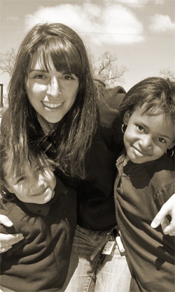 A woman smiling and embracing two young girls outdoors. All are wearing dark jackets, and the background shows a clear sky with a few clouds. The image has a warm, sepia tone.