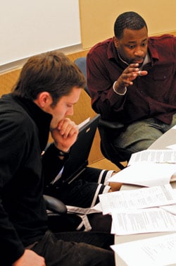Two men sit at a table covered with papers. One man gestures while speaking, and the other listens, resting his chin on his hand. A laptop is open in front of them. They appear to be engaged in a discussion or work session.