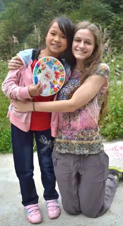 A young girl in a pink jacket and jeans holds a colorful paper plate while standing next to a smiling woman with long hair. They are outdoors, surrounded by greenery. The woman kneels beside the girl with her arm around her.