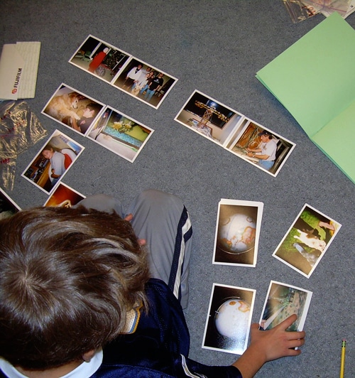 A child sits on the floor arranging photos in a semicircle. An open green folder is nearby, and a pencil lies on the carpet. The photos display various scenes and people.