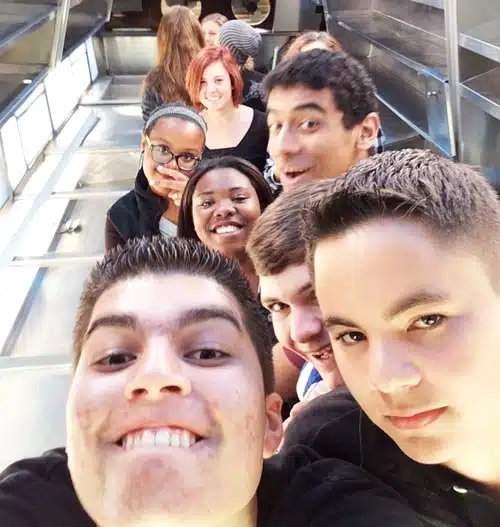A group of smiling and laughing teenagers takes a selfie on an escalator. They are casually dressed, and the atmosphere is lively and fun. The background shows the metal and glass structure of the escalator.