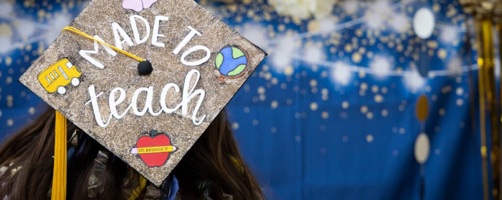 A graduation cap decorated with glitter, a bus, globe, heart, apple, and the words Made to teach against a festive, starry blue background celebrates the San Diego Teacher Residency (SDTR) journey.