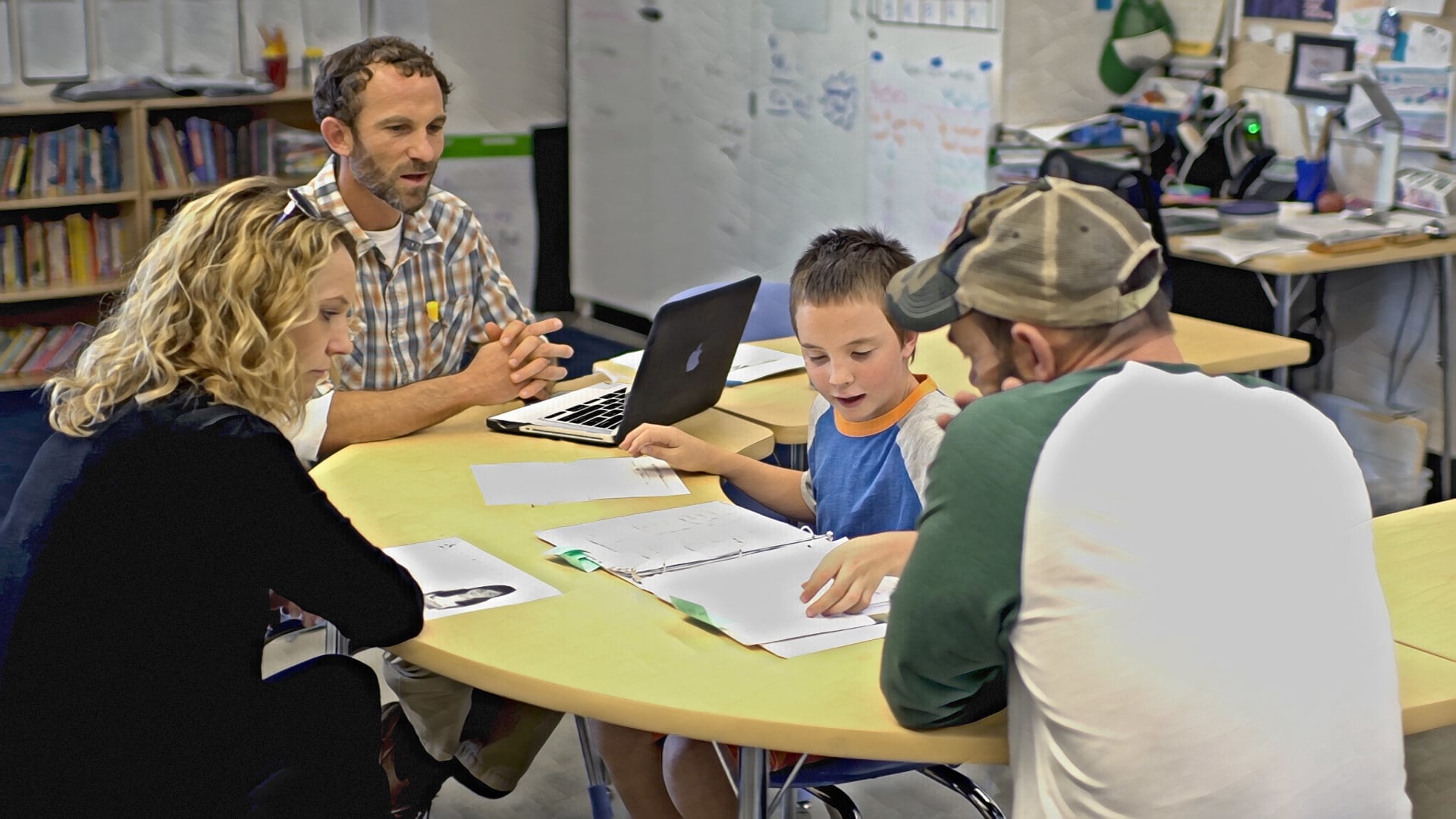 A young boy shows his work in a binder to three adults seated around a table in a classroom. One adult has a laptop open. Bookshelves, a whiteboard, and educational materials are visible in the background.