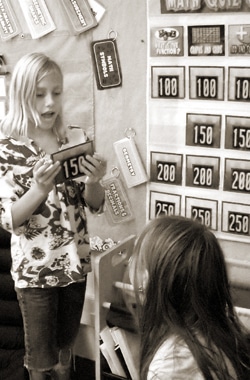 A child holds a card displaying the number 150 in front of a wall with quiz scores. Another child is seated, looking up at them. Both are engaged in a discussion or activity, possibly related to a math quiz. The image is in black and white.