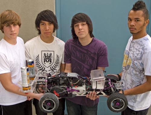 Four young people standing side by side, holding a large remote-controlled car. They are dressed casually and appear to be in an indoor setting with a blue door behind them. The car is detailed, showing various components and wheels.
