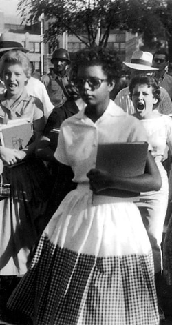 A young Black woman walks holding books, her expression focused. Behind her, a crowd follows, with a young white woman appearing to shout. The scene conveys tension and determination.