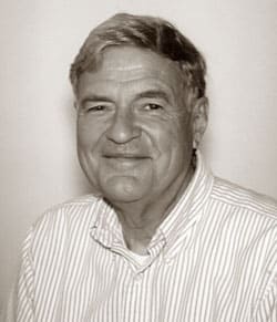 A sepia-toned portrait of an older man with short, light-colored hair. He is smiling and wearing a striped button-up shirt, looking directly at the camera.