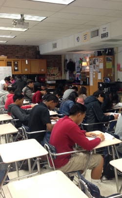A classroom filled with students sitting at desks. They are focused on writing or taking an exam. The room has various educational posters and materials on the walls, and shelves at the back. The setting suggests a busy and studious environment.
