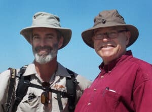 Two men standing side by side outdoors under a clear blue sky. Both wear wide-brimmed hats and smiles. The man on the left has a beard and backpack, while the man on the right wears glasses and a red shirt.