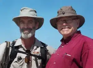 Two men standing side by side outdoors under a clear blue sky. Both wear wide-brimmed hats and smiles. The man on the left has a beard and backpack, while the man on the right wears glasses and a red shirt.