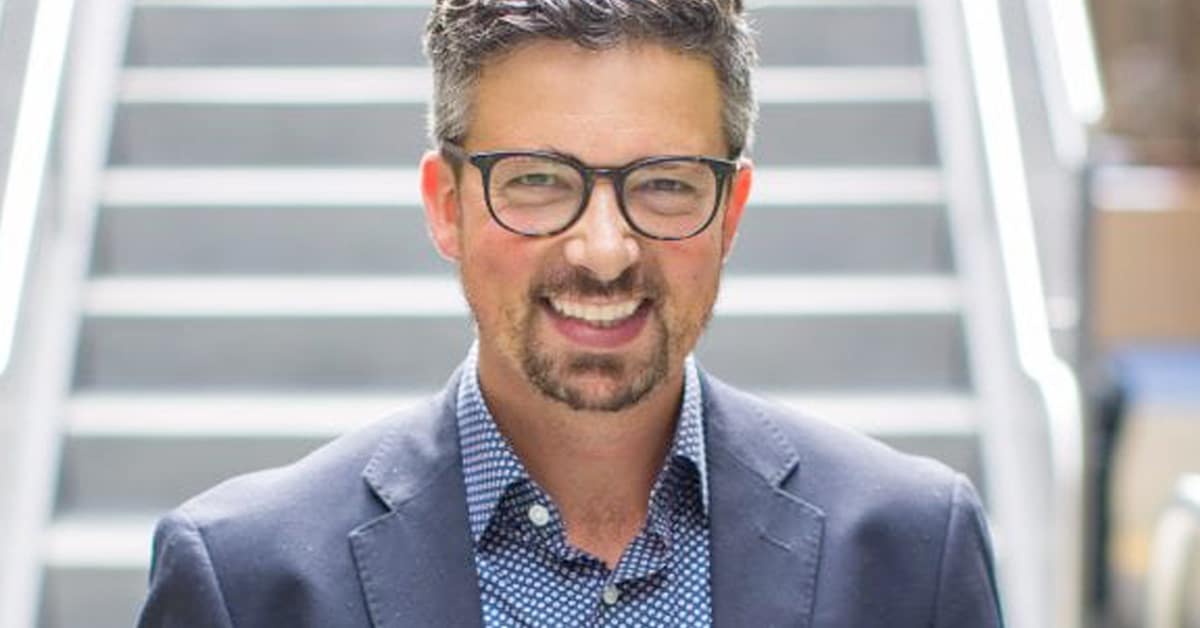 A man with short hair, wearing glasses, a dark blazer, and a patterned shirt, smiles in front of a set of stairs.