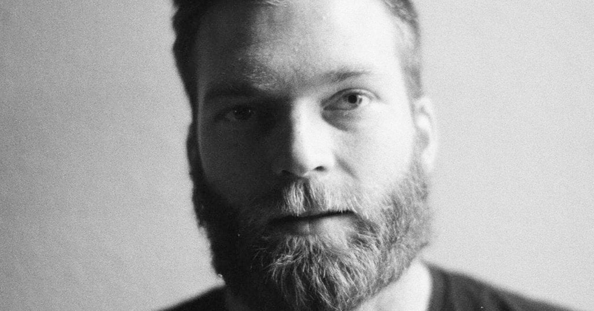 Black and white close-up portrait of Brent Spirnak, a man with a full beard and mustache. He has short hair and is looking directly at the camera with a neutral expression. The background is plain and softly blurred.
