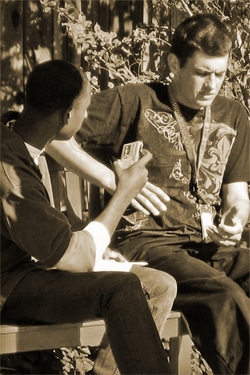 Two men sit on a bench outdoors. One man holds a phone, showing it to the other. They appear to be engaged in a discussion. The image has a sepia tone, giving it a vintage look.