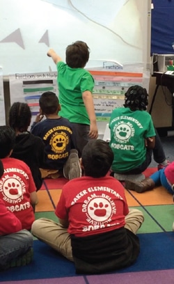 Children sit on a colorful rug, wearing matching shirts with Baker Elementary on the back. One child stands, pointing at a chart titled Fractions displayed at the front of the classroom.