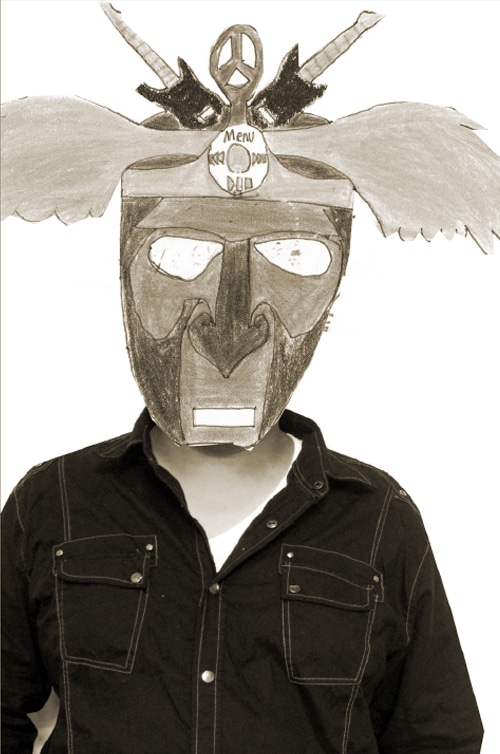 A person wearing a detailed, hand-crafted mask with feathers, peace symbols, and decorative elements, posing in a dark shirt. The mask is large, covering the entire face, and is set against a simple background.