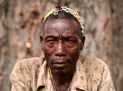 A man wearing a patterned headband and a beige shirt is looking at the camera. The background is a tree bark.