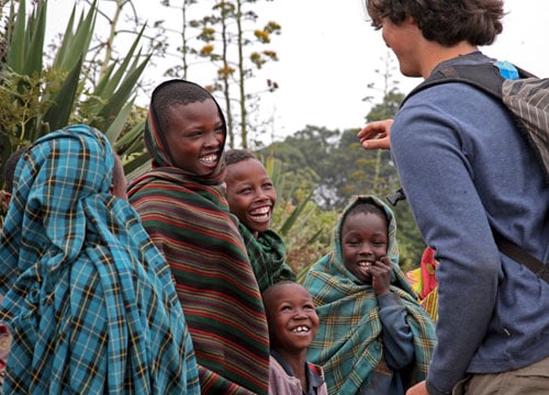 A group of children wrapped in colorful blankets smile and laugh outdoors, engaging with a person wearing a blue shirt and a backpack. There is greenery in the background, creating a lively and cheerful scene.