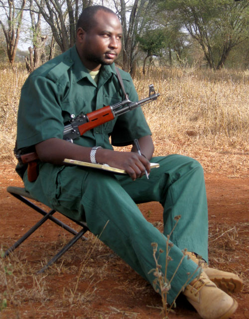 A man in green work clothes sits on a folding chair in a dry, grassy landscape, holding a pen and notepad. An automatic rifle is slung over his shoulder, and he wears tan boots. Sparse trees are visible in the background.