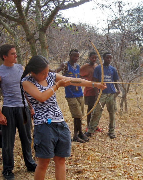 A woman in a zebra-striped shirt practices archery with a bow in a dry, wooded area, watched by four men. Three of the men stand in a line behind her, and one stands to her side, observing closely.