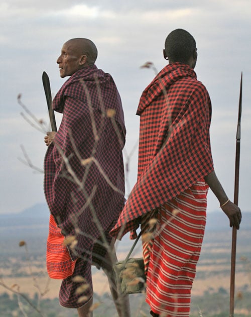Two people dressed in traditional Maasai attire stand on a hill, holding spears. They overlook a vast landscape under a cloudy sky, with one wearing a red checkered shawl and the other a striped one. Sparse vegetation is visible in the foreground.