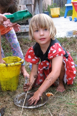 A child in a red polka-dot dress plays with mud outdoors, using a bucket and a metal tray. She is barefoot and appears engaged in the activity, with a playground and toys visible in the background.