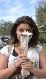 A person holding a large bone in front of their face, standing outdoors. The bone covers part of their face, and theyre surrounded by trees and a clear sky.
