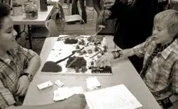 Two kids play a board game on a table covered with various pieces. They are focused and engaged in the activity. The background shows other people and chairs in a room, suggesting a group event or gathering. The image is in black and white.