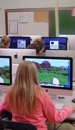 Students in a classroom sit at desks using computers, playing Minecraft. The screens show a virtual landscape. The classroom has white walls and a green chalkboard.