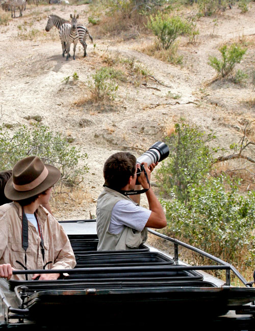 Two people on a safari in an open-top vehicle. One is photographing zebras in the distance with a large camera lens, while the other looks on. The landscape is dry and rugged with sparse vegetation.