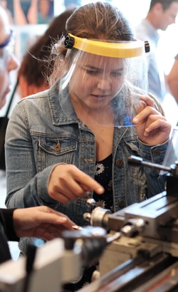 A person wearing a denim jacket and a face shield operates a lathe machine. They are focused on the task, with another person nearby assisting or observing. The background is slightly blurred.