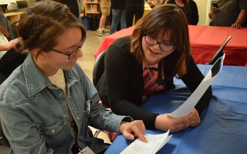 Two women sitting at a table covered with a blue cloth, engaged in conversation while looking at documents. They are surrounded by people and a background of bookshelves.