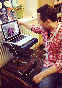 A person in a checkered shirt is kneeling next to a makeshift laptop stand made of stacked books. The laptop screen shows a video call with another person. The scene appears to be in a library or study area.