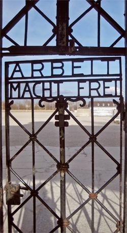A black iron gate with the inscription ARBEIT MACHT FREI viewed from the inside, opening to an empty paved area and distant buildings under a blue sky.