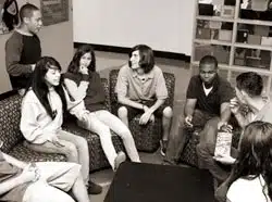 A group of teenagers sit together on couches and chairs, engaging in conversation. They are indoors, and one person is gesturing while speaking. The setting has a casual, relaxed atmosphere.