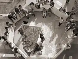 Aerial view of a group of people gathered around an octagonal structure on a tiled surface. Some are sitting on the structures ledges, while others are standing. A large tarp is spread out in the center.