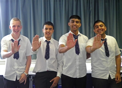 Four young men in school uniforms stand in a row, smiling and holding their right hands up. They are wearing white shirts and ties, and a curtain hangs in the background.