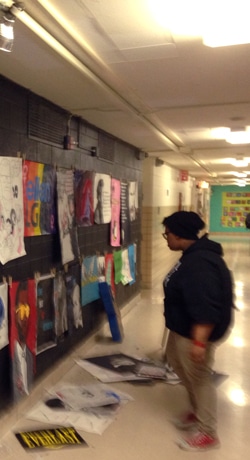A person wearing a beanie and hoodie observes artwork displayed on a school hallway wall. Various colorful and monochrome posters and drawings are attached to the wall, with some spilling onto the floor.