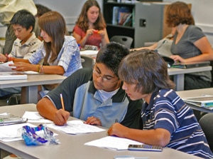 Students are engaged in group work at a classroom table. Two boys in the foreground are focused on a worksheet, while other students are seated in the background, discussing and writing notes. The room is bright and organized.