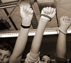 Three children raise their right arms and fists in the air, wearing wristbands. They appear to be in a room with visible ceiling pipes and light fixtures. The photo is in black and white.