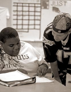A student and a teacher are engaged in a discussion over a notebook. The student is seated, wearing a white t-shirt, and the teacher is leaning in, wearing a sports jersey and cap. A classroom board is visible in the background.