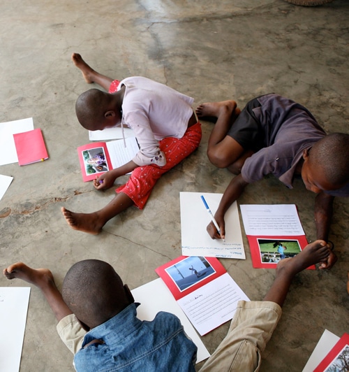 Three children are sitting on the floor, writing in notebooks. Papers with photos are scattered around them. They are focused on their work, and the floor is concrete.