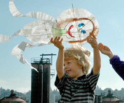 A smiling child with curly blond hair is flying a hand-painted kite with a face design. The kite has blue eyes and a red smile. Another person helps hold the kite string. Industrial structures are visible in the background under a clear sky.