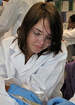 A young woman in a lab coat, safety goggles, and blue gloves is focusing intently on a task, possibly a science experiment. She is surrounded by lab equipment and colleagues in similar attire.