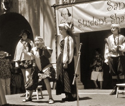 A sepia-toned image of four people in Shakespearean costumes performing on a stage. One person is sitting on a chair, gesturing expressively, while others stand nearby. A banner above reads, Student Shakespeare Festival.