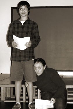 A young person stands on a stage holding papers, smiling. Next to them, an adult kneels, looking up, also holding papers. The background features a blank screen.