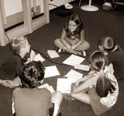 A group of five children sit in a circle on the floor, engaging in a discussion. Papers are spread out in the center. The children appear to be in a classroom or similar setting. The image is in black and white.