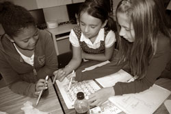 Three young students collaborating on a school project at a desk, surrounded by notebooks and pens. The image is in black and white, highlighting their focused expressions and teamwork.