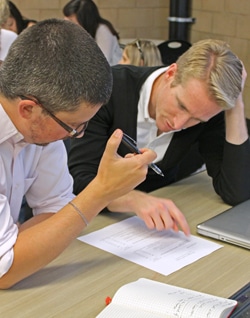 Two men sitting at a table, discussing a document in front of them. One wears glasses and gestures with a pen, while the other leans forward, focused on the paper. A laptop and a notebook are also on the table.