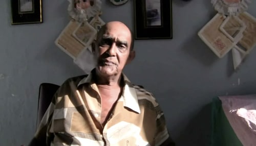 An elderly man wearing a patterned shirt sits in a dimly lit room. Behind him, there are picture frames and decorative items on the wall.